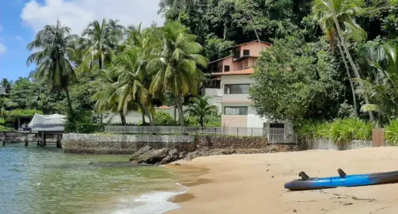 Casa de temporada vista da praia. Em volta da casa há uma vegetação com árvores e coqueiros e no lado esquerdo está o mar. Imagem para ilustrar o post airbnb em Angra dos Reis.