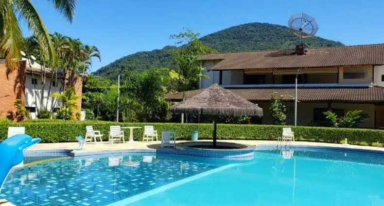 Foto da área externa da Casa Melo 4 para ilustrar post sobre airbnb em Perequê Mirim. Uma piscina com uma ilha de quiosque e cadeiras de praia ao redor. Ao redor um jardim e no fundo prédios