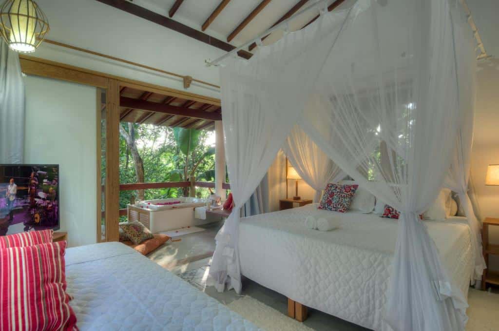 Um dos quartos do Casas Natureza Brasil. Uma cama de casal com dossel está no meio, de frente para outra cama menor, de solteiro. Ao fundo é possível ver uma banheira coberta por pétalas de rosa, de frente para a visão do jardim. Este também é um airbnb em Arraial d'Ajuda.