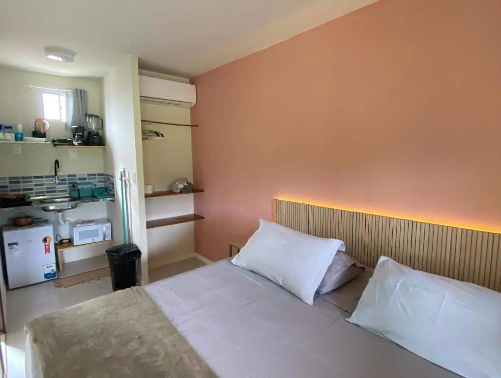 Quarto da CASA PITANGA. Do lado direito uma cama de casal, no fundo, no lado esquerdo um frigobar, um micro-ondas e uma pia com utensílios da cozinha. Do lado direito prateleiras e em cima um ar-condicionado. Foto para ilustrar post sobre airbnb em Fernando de Noronha.