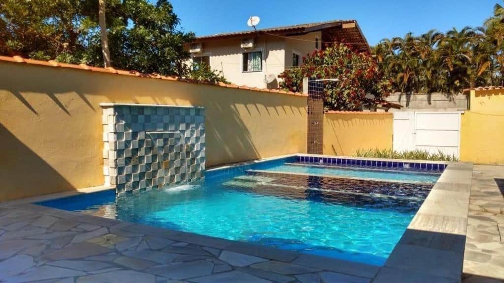 Área livre da Casa Temporada. Uma piscina no meio com cascata, no fundo o portão da casa e uma ducha.