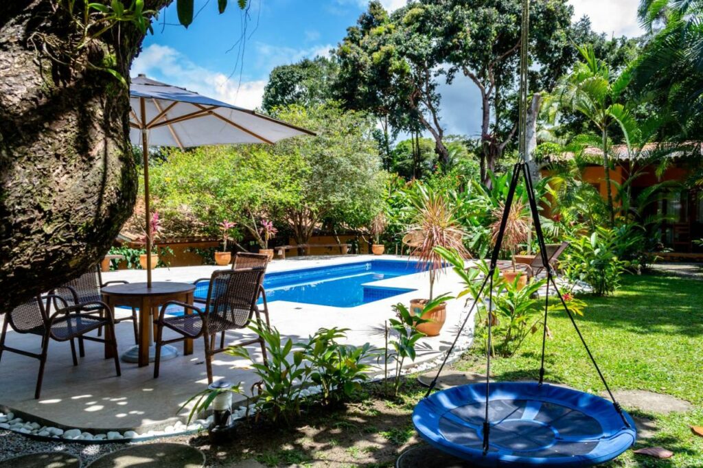 Área externa do Casas Natureza Brasil. Uma pisicna está ao fundo, em um espaço de piso. Perto dela há uma mesa de vários lugares com um guarda-sol. Ao redor o espaço é todo de grama, cheio de plantas e com um balanço.