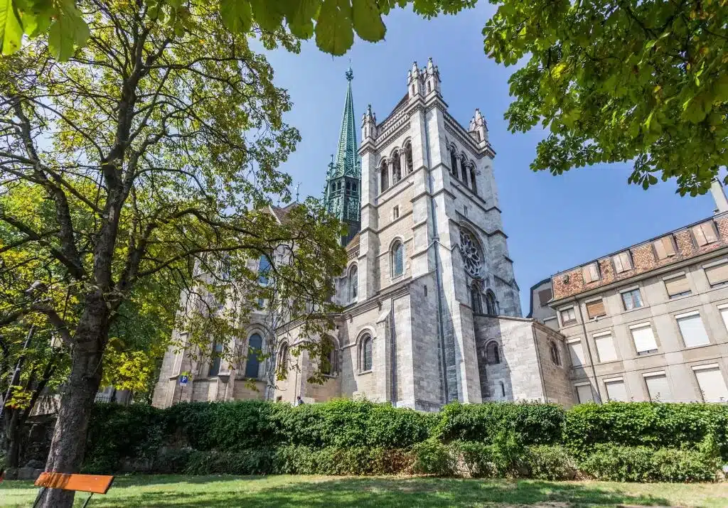 Imagem da Catedral St. Pierre, em Genebra. Está de dia e vemos a alta construção da igreja, a sua volta há vários arbustos e árvores.