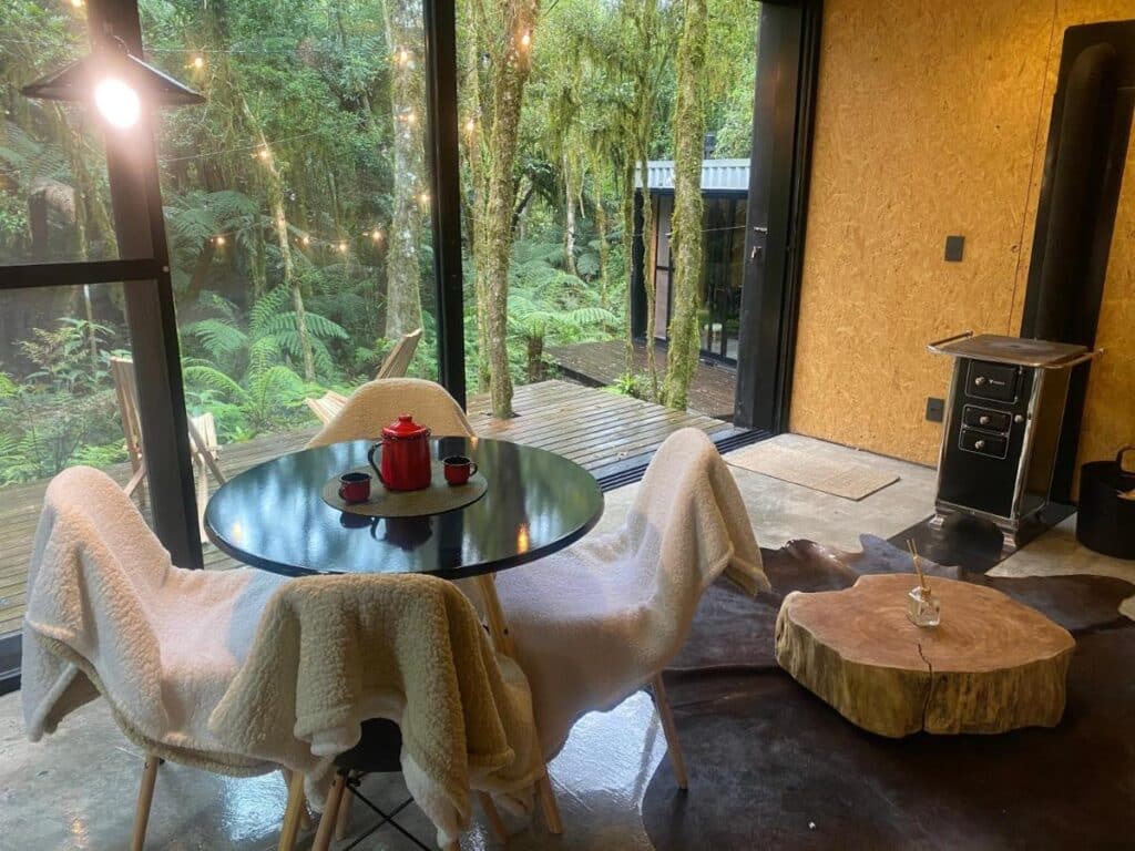 Uma mesa com cadeiras no Chalé dos Alpes. Do lado direito uma lareira. No fundo uma parede de vidro.