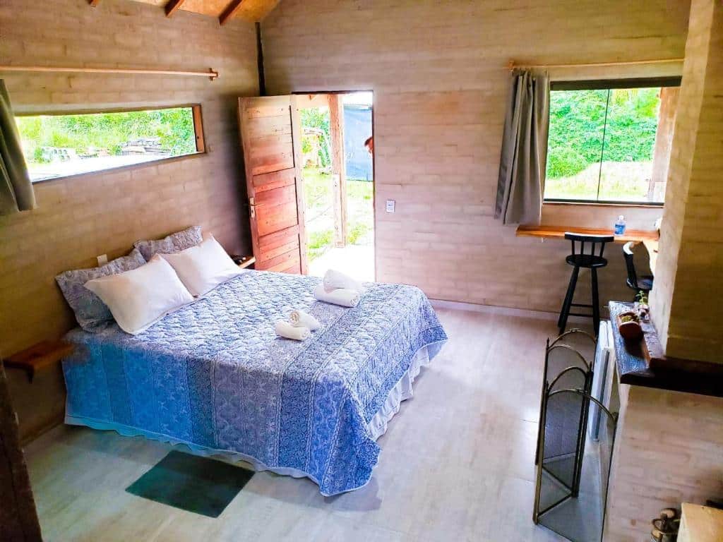 Quarto do Chalé na Montanha. Do lado esquerdo uma cama de casal, no fundo a porta do quarto. Do lado direito uma lareira, no fundo uma bancada com bancos e a janela. Foto para ilustrar post sobre airbnb em Santo Antônio do Pinhal.