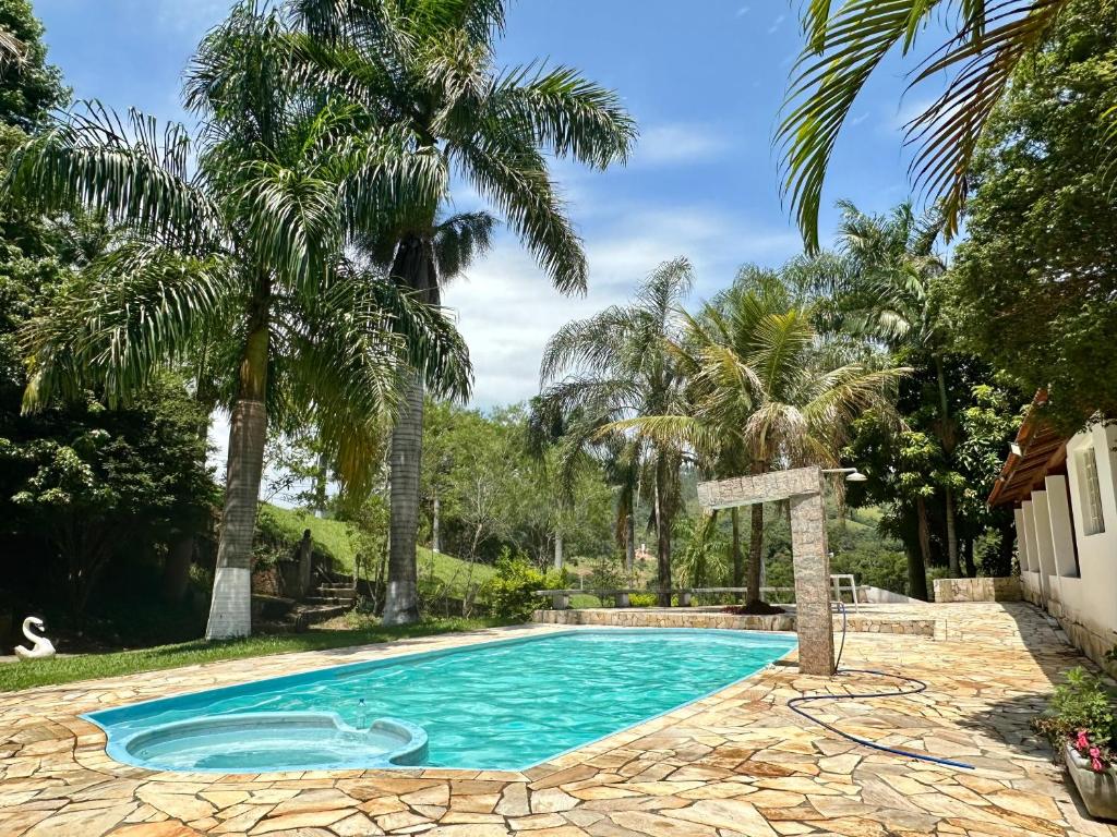 Uma piscina com deck de pedra cercada por palmeiras e vegetação em um dia ensolarado, na propriedade do Chalé Recanto Luz da Montanha.