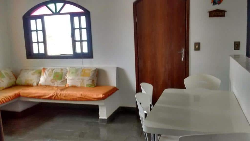 Cozinha do Chalés Monções. Do lado direito uma mesa com cadeiras, no fundo a porta. Do lado esquerdo um sofá cama, em cima uma janela.
