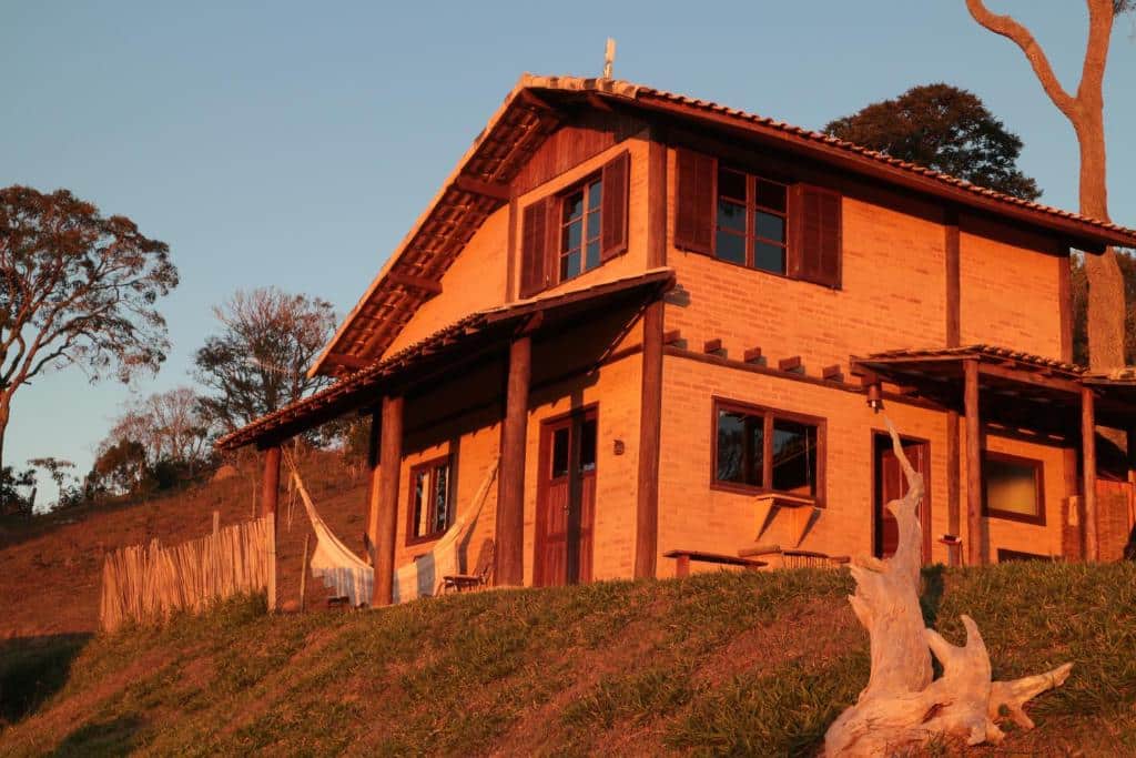 Um dos airbnb dos Chalés Samadhi que mostra uma casa de dois andares em formato de chalé com paredes de tijolos e janelas, portas e detalhes em madeira. Na varanda da casa tem uma rede, algumas árvores em volta e um gramado verde. Imagem ilustrando post Airbnb em Cunha.
