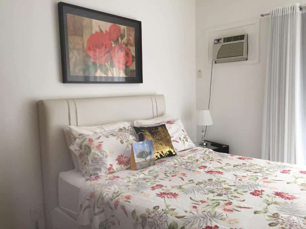 Quarto do airbnb Charme Comforto Beira Mar. A cama está encostada na parede do lado esquerdo do quarto, no lado direito da cama há uma pequena cômoda com um abajur em cima e ainda no lado direito é possível ver um pouco de uma cortina. Imagem para ilustrar o post airbnb em Angra dos Reis.