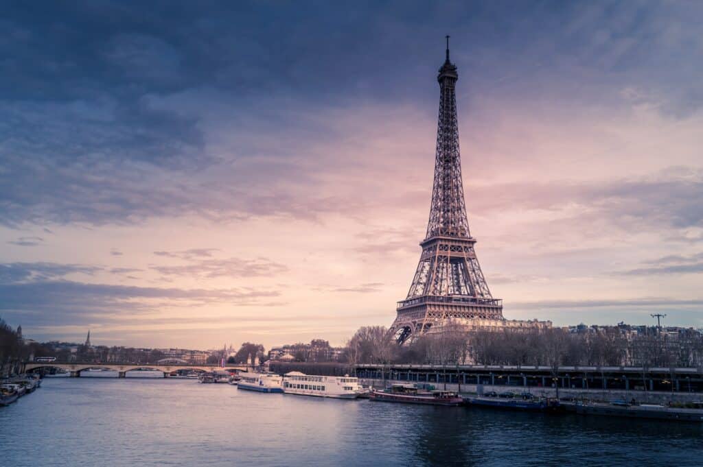 A Torre Eiffel do lado direito da foto com um rio passando ao lado com barcos estacionados e uma ponte, além de algumas árvores às margens do rio