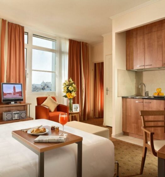 Quarto do Citadines Saint-Germain-des-Prés Paris, um dos hotéis com cozinha em Paris. Uma cama de casal tem uma mesinha com café da manhã logo ao lado e encara a cozinha em frente, que é equipada com pia, micro-ondas, cooktop e frigobar. Ao lado direito da cama fica uma mesa de trabalho com cadeira, e ao lado esquerdo há um suporte com TV e aparelho de som, para representar hotéis com cozinha em Paris