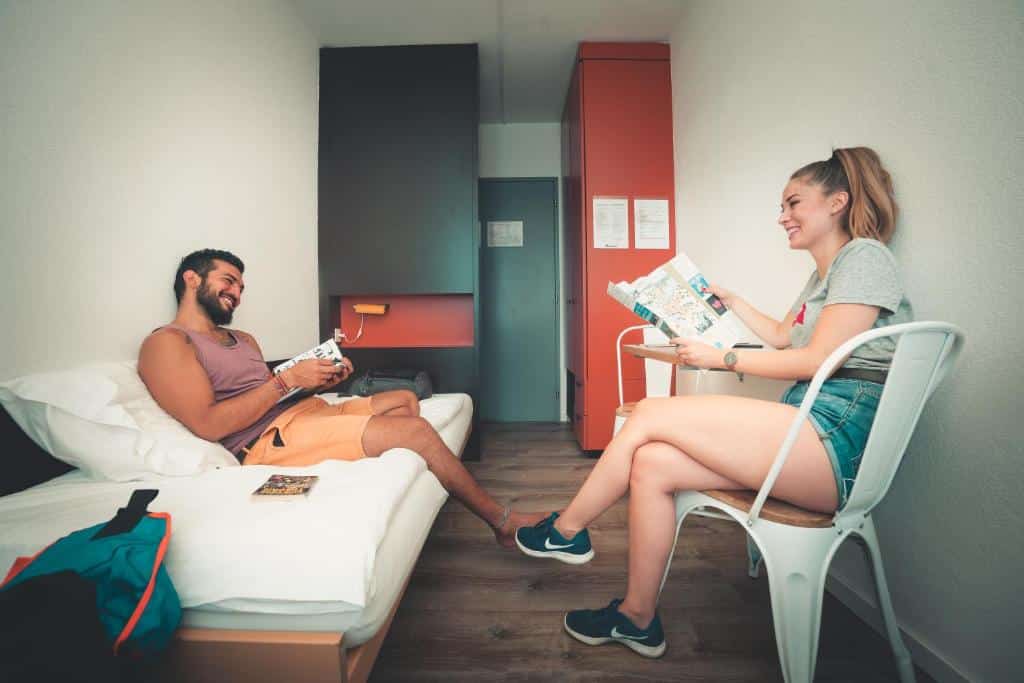 Uma moça loira, com um guia de viagem na mão, sentada em uma cadeira branca na direita de uma suíte no City Hostel Geneva, conversando com um rapaz bronzeado. O homem está sentado na cama à esquerda, com um livro na mão.