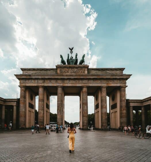 Um monumento com quatro colunas, no alto há uma escultura com cavalos e homens, em uma praça com pessoas andando, o local é conhecido como Pariser Platz 1, para representar o que fazer em Berlim