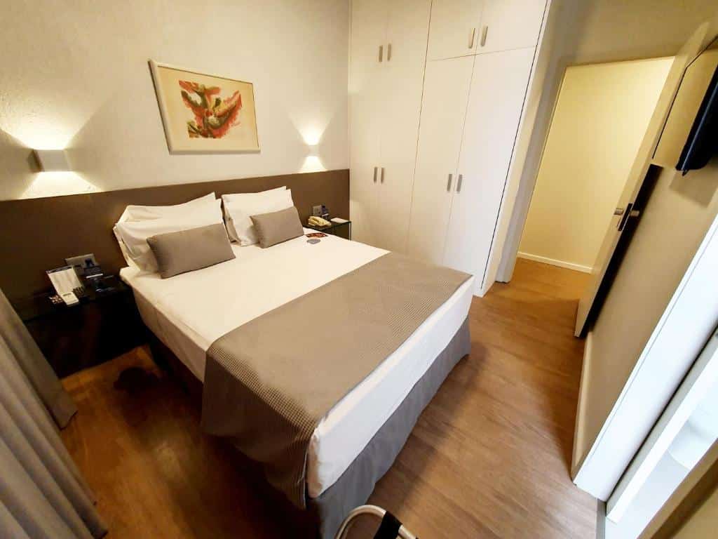 Foto de cima do quarto em Max Savassi Apart Service. A cama é box de casal, está no meio do ambiente. Há uma mesa de cabeceira em seus dois lados. Na direita, há um guarda-roupa e porta para o quarto. Na frente da cama uma TV e porta para o banheiro. Representa o post de airbnb em Belo Horizonte.