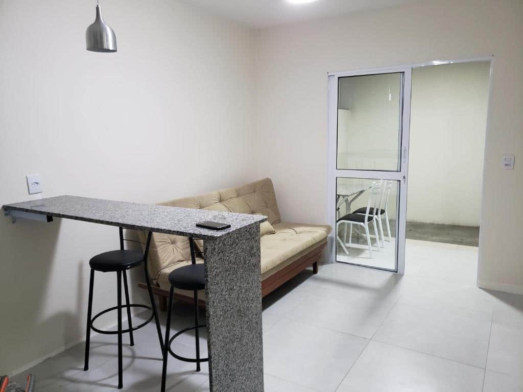 Sala de estar do Condomínio Sophia Silva. Do lado direito um calcão com bancos, no fundo um sofá cama, no fundo a varanda com mesa e cadeiras.