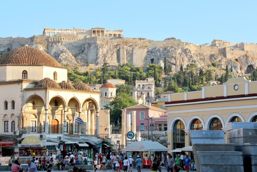 Bairro de Monastiraki em Atenas, a região conta com comercial ao ar livre, pessoas caminhando e, mais ao fundo, prédios históricos, além das ruínas