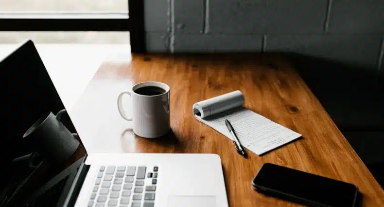Foto tirada na lateral de uma mesa com um notebook aberto. Ao lado há uma caneca de café, um bloco de notas com uma caneta em cima e um celular. No cento superior esquerdo, uma janela ilumina o ambiente. A imagem ilustra o post sobre como comparar seguro viagem. - Foto: Andrew Neel via Unsplash