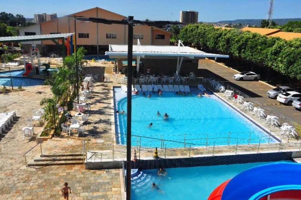 Área externa do diRoma Fiori - Apartamentos JN. Duas piscinas do lado direito, com pessoas e cadeiras de praia ao redor. Na frente a ponta de um escorregador aquático. Do lado esquerdo uma piscina infantil com brinquedos.