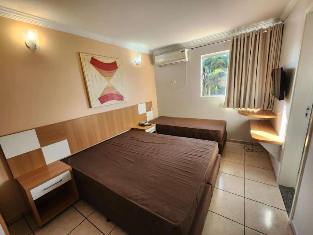 Quarto do diRoma Fiori - Apartamentos JN. Uma cama de casal e uma cama de solteiro do lado esquerdo, entre elas uma cômoda, no fundo em cima um ar-condicionado e uma janela com cortina. Do lado direito duas prateleiras, uma televisão e uma porta. Foto para ilustrar post sobre airbnb em Caldas Novas.
