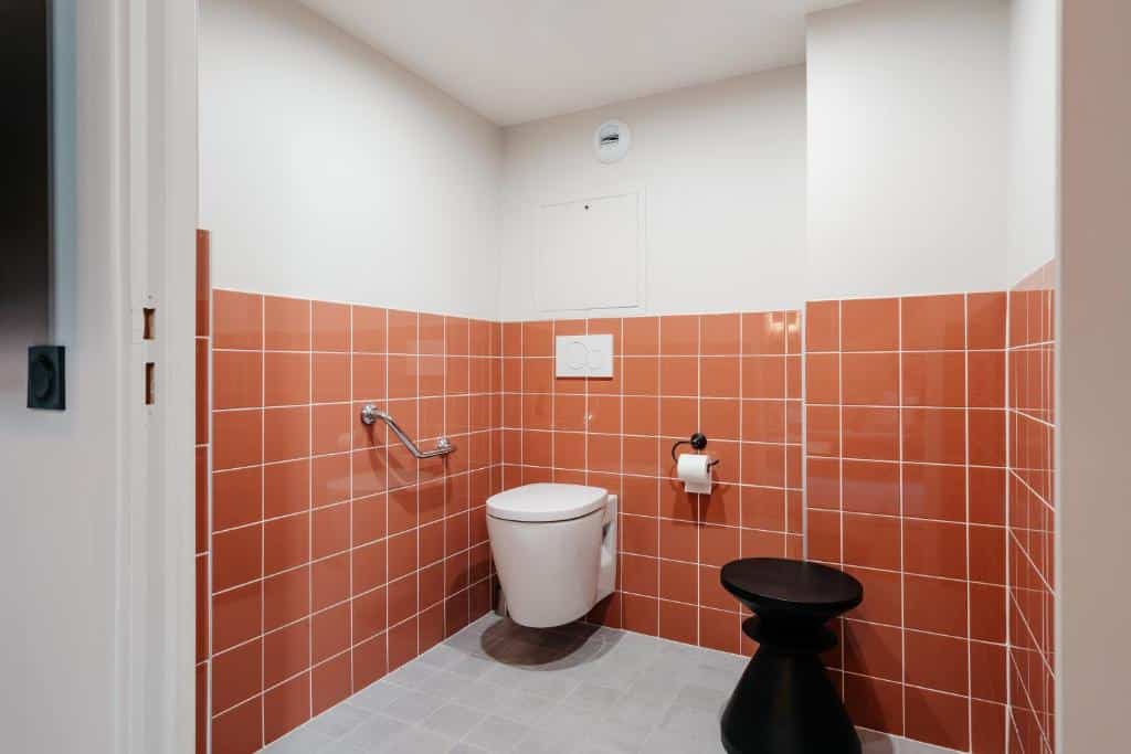 Banheiro adaptado para PCD no Edgar Suites Louvre - Jour. Uma privada elevada tem uma barra de apoio na parede esquerda e um rolo de papel higiênico ao lado direito. Há também um banco preto ao lado do papel.