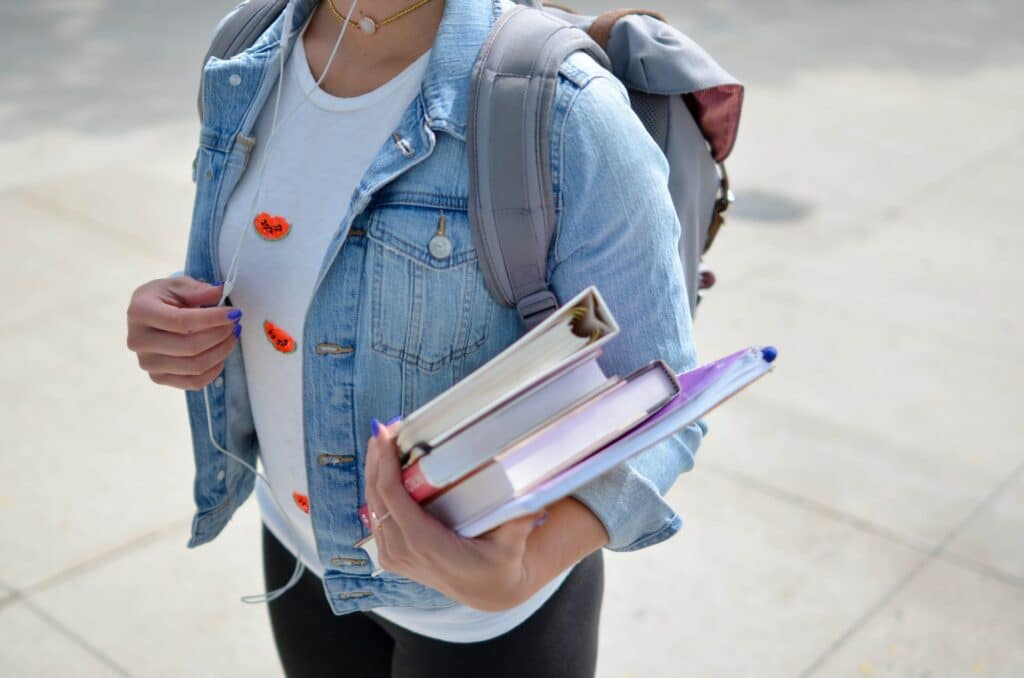Uma menina usando uma jaqueta jeans e uma mochila nas costas, em um dos ante-braços ela está apoiando livros e uma pasta, para representar seguro viagem Irlanda