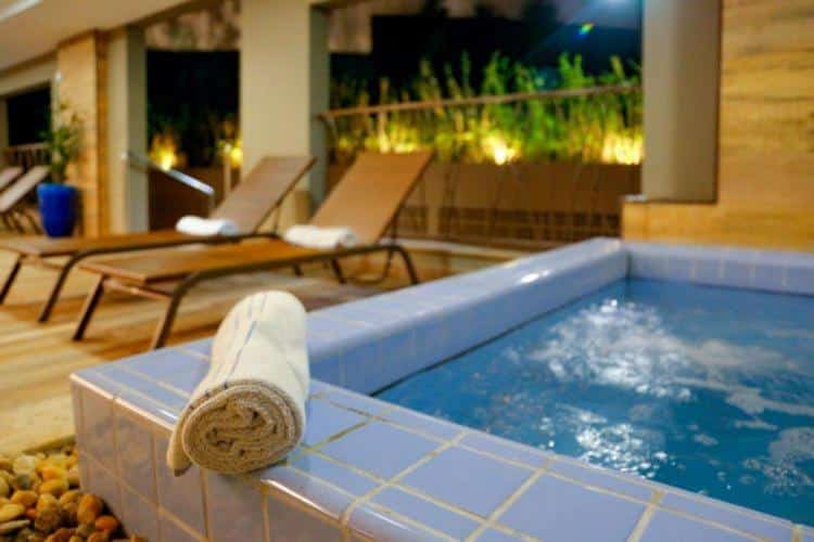 Piscina do Enjoy Olimpia Park Resort. Do lado direito a piscina, no meio uma toalha enrolada, no fundo cadeiras de tomar sol.