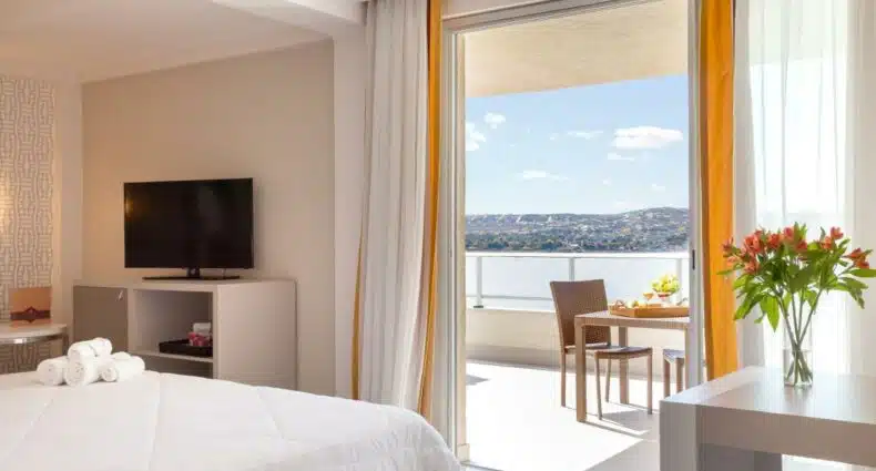 Quarto do hotel eSuites Spa, em Lagoa Santa. A cama está virada para a porta da varanda, que está aberta e mostrando uma mesa com cadeira de frente para a vista do lago. Essa é uma opção na lista de hotéis e pousadas em Lagoa Santa.