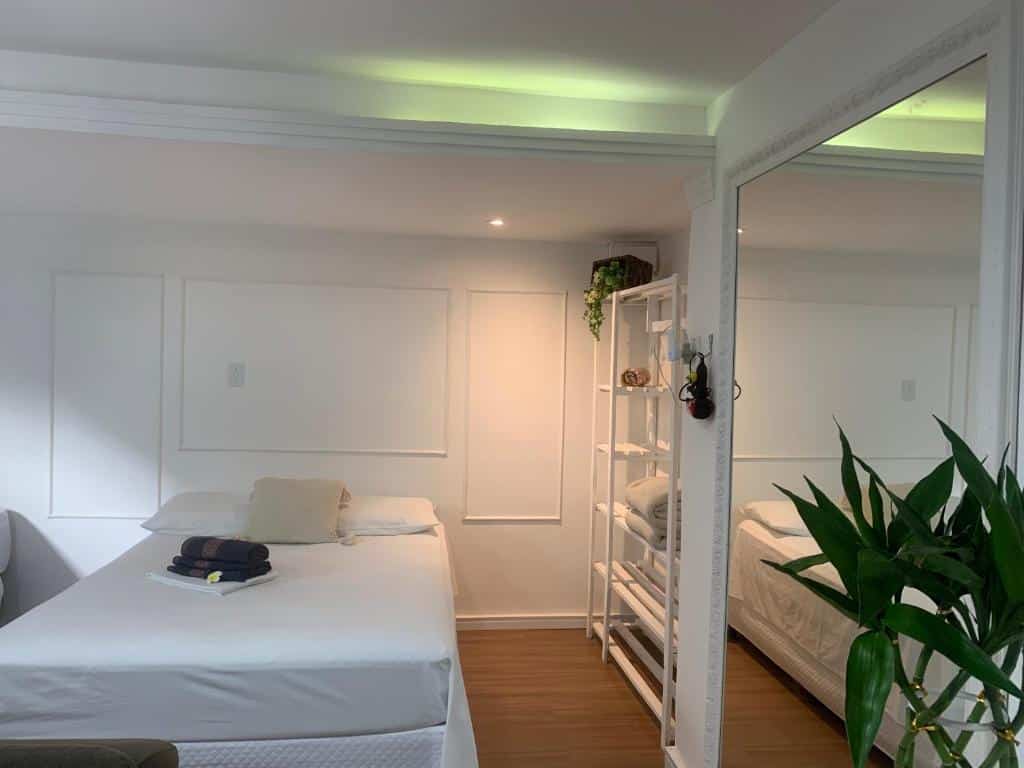 Quarto do LOFT BC. Do lado direito uma planta, um espelho e uma estante. No meio uma cama de casal com toalhas e travesseiros em cima.