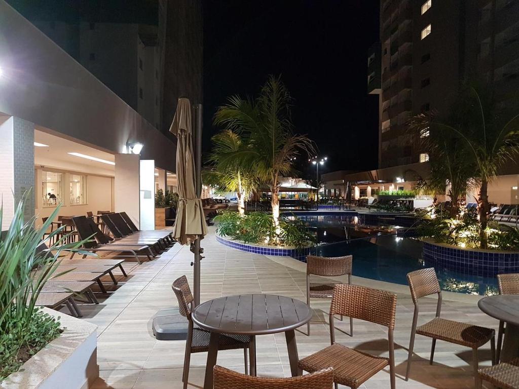 Área externa do Flat Olímpia Park Resort. Mesas, cadeiras e cadeiras de tomar sol, no meio uma piscina com palmeiras nos cantos.