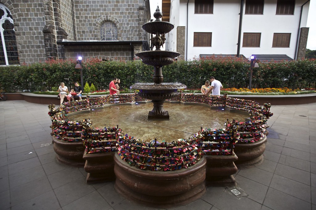 Fonte do Amor Eterno em Gramado circular cercada por grades de metal cobertas com vários cadeados, com pessoas sentadas nas bordas ao fundo.