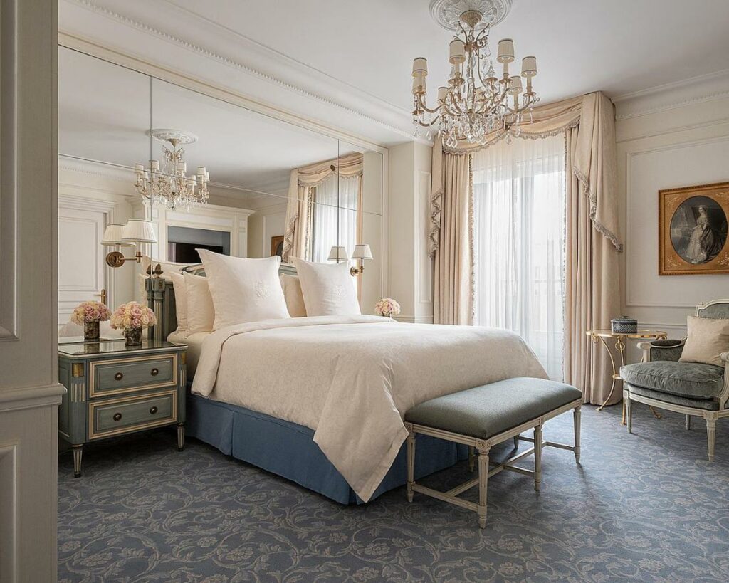 Quarto do Four Seasons Hotel George V Paris. A cama de casal tem um banco acolchoado aos pés e mesinhas de cabeceira com vaso de flores e abajur dos dois lados. A parede atrás da cama é feita de espelhos. Ao lado direito da cama há uma janela com cortinas fechadas, e uma poltrona e uma mesinha ficam logo ao lado. Um lindo lustre está pendurado acima da cama.