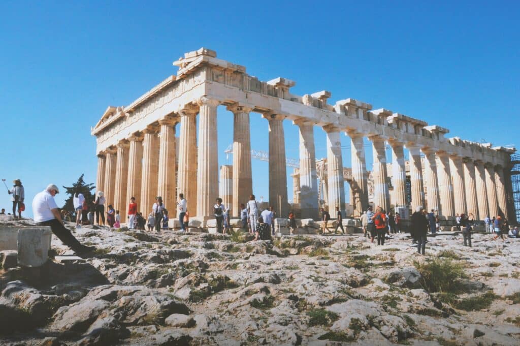 Acrópole de Atenas, uma rúina com diversas colunas em tons de bege claro, com pedras ao redor e diversas pessoas caminhando no local