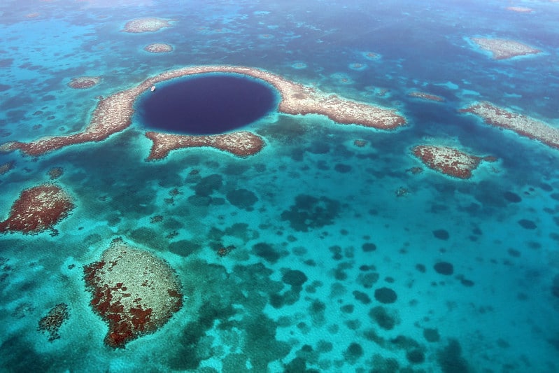 Imagem aérea de um destino turístico em Belize, o Grande Buraco Azul. Um mar de águas cristalinas com vários corais e uma profundidade circular no meio.