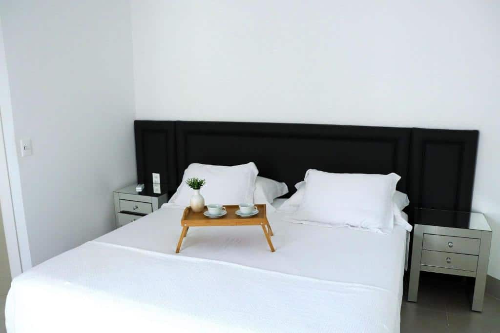 Quarto do Haus Stay Pé na Areia. Uma cama de casal com uma cômoda de cada lado e uma mesinha de madeira com xícaras e um vaso de flor em cima da cama. Foto para ilustrar post sobre airbnb em Balneário Camboriú.