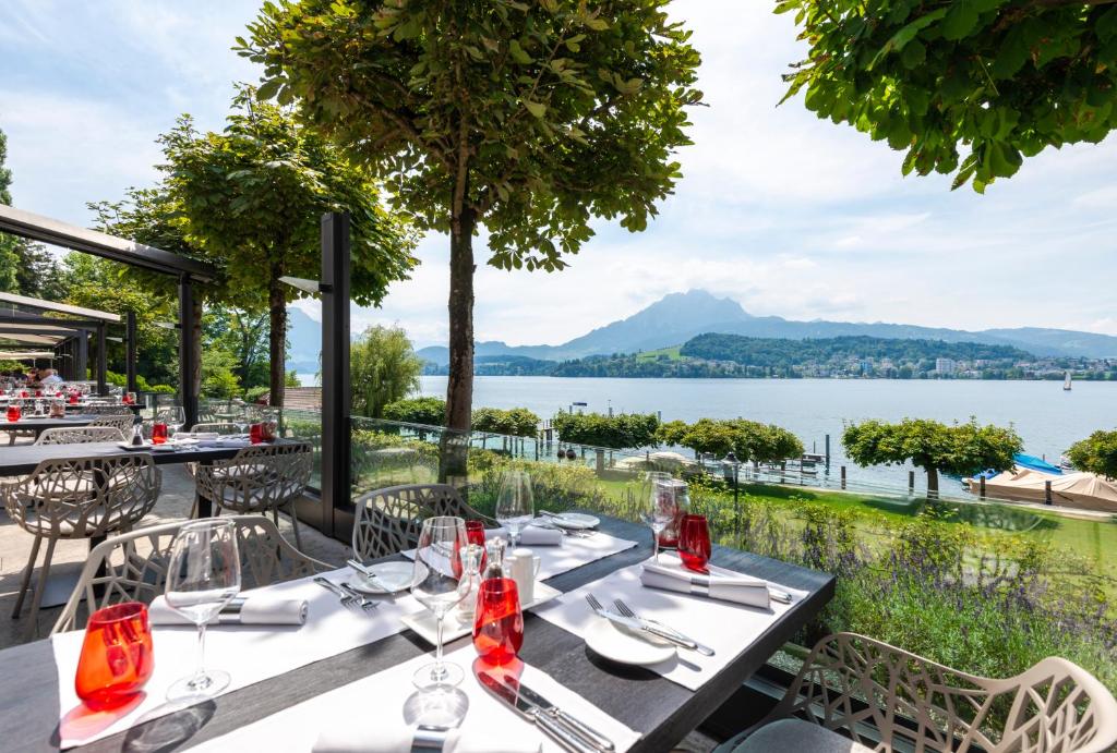 Restaurante ao ar livre no Hermitage Lake Lucern. Na esquerda, em cima e um deck, vemos mesas de madeira preta com cadeiras e talheres, copos e guardanapos em cima. Dele, há uma vista para a natureza em volta, que consiste em um jardim, um lago ao fundo e montanhas no horizonte.