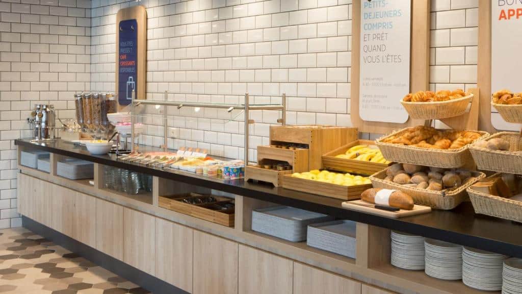 Buffet do Holiday Inn Express Geneva Airport. A bancada é extensa e ocupa a parede inteira. Há muitas opções de pães, frutas, cereais, sucos, etc. Vemos embaixo, em compartimentos, pratos e bandejas.