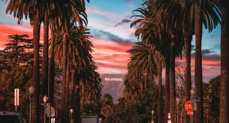 Placa de Hollywood vista de longe. Em primeiro plano estão as diversas árvores na rua de Beverly Hills. O céu está colorido, como se estivesse no final do dia. Essa foto ilustra o post de o que fazer em Los Angeles.