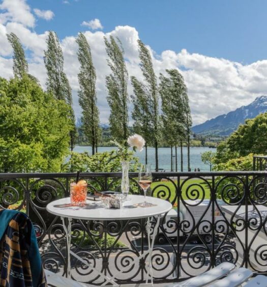 Vista de uma varanda no Hotel Beau Séjour. Há uma mesa com duas cadeiras perto da grande, com copos e taças em cima. A vista é de um belo dia com o céu azul e com nuvens. Há árvores sendo sopradas pelo vendo, um lago e montanhas ao fundo.