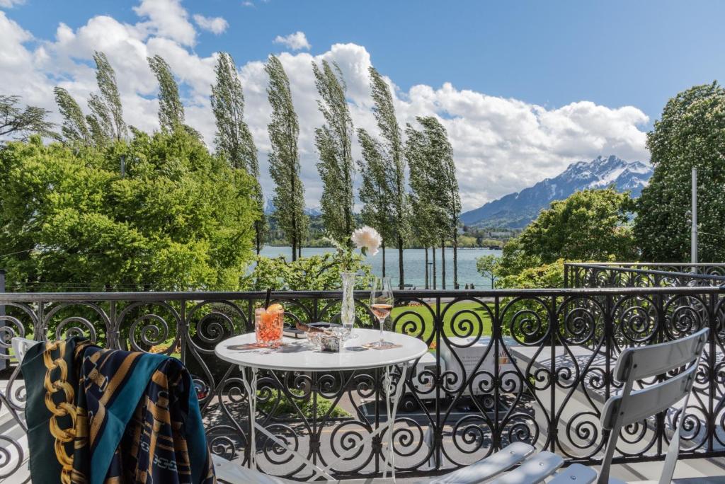 Vista de uma varanda no Hotel Beau Séjour. Há uma mesa com duas cadeiras perto da grande, com copos e taças em cima. A vista é de um belo dia com o céu azul e com nuvens. Há árvores sendo sopradas pelo vendo, um lago e montanhas ao fundo.