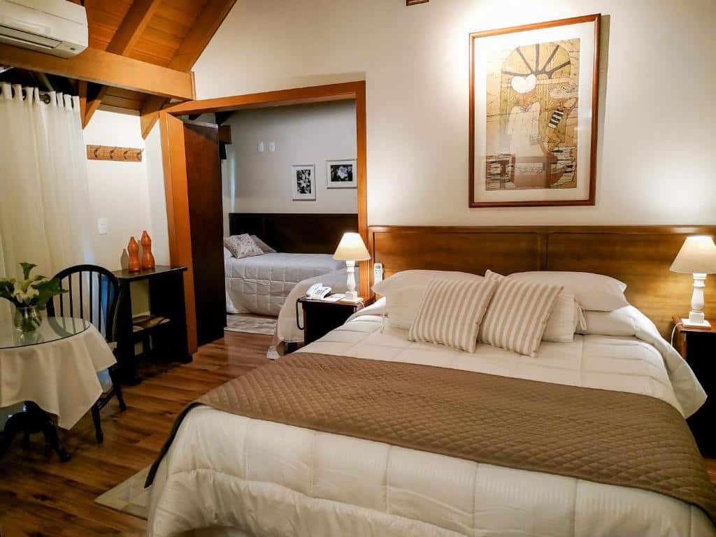 Quarto do Hotel Canto Verde para ilustrar post sobre hotéis baratos em Gramado. Uma cama de casal do lado direito, com uma cômoda e um abajur de cada lado. Do lado esquerdo uma mesa com duas cadeiras e uma estante. No fundo, uma porta aberta com uma cama de solteiro.