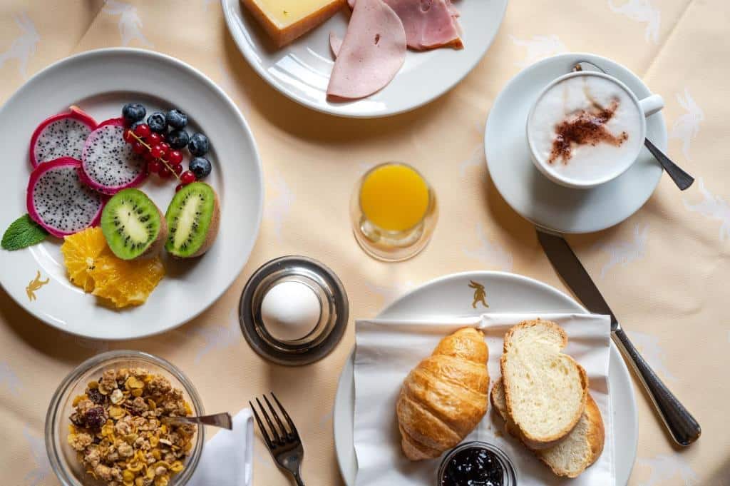 Exemplo de um café da manhã no Hotel De la Paix. A mesa possui pratos com pães, croassaints, frutas, geleias, queijos e frios Há uma xícara com cappuccino e uma tigela com cereais.