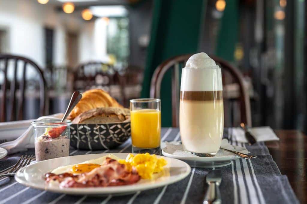 Exemplo de um café da manhã no Hotel Hofgarten Luzern. Vemos um prato com ovos e bacon, um copo com frappé e outro com suco, iogurte e uma cesta com pães e massas.