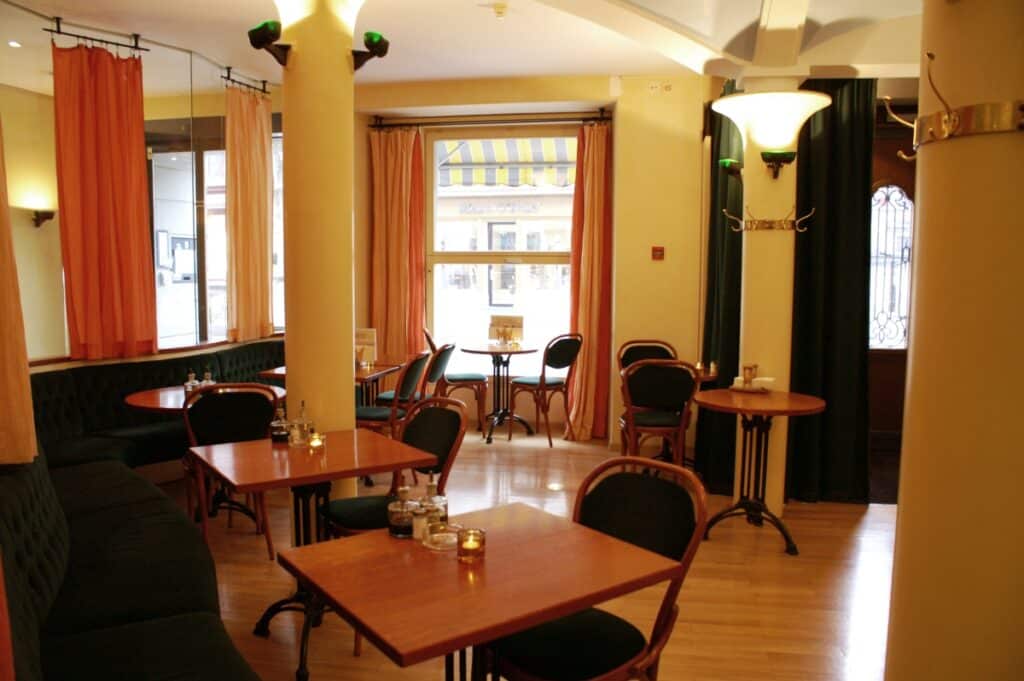 Foto do restaurante no Altstadt Hotel Krone. O piso é de madeira clara, e as mesas também, acompanhadas de duas cadeiras cada Há colunas no ambiente e alguns lustres. Janelas altas com cortinas laranjas também estão presentes.