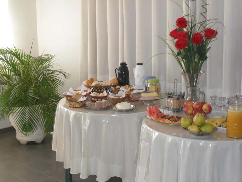 Mesas de café da manhã do Hotel Lund. São duas mesas redondas com frutas, frios, pães, queiro e algumas outras coisas. Um vaso de flor decorativo também está ali.