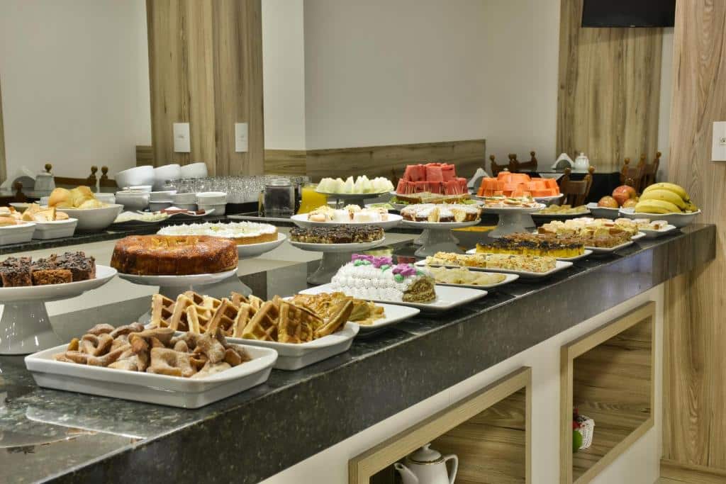 Bancada do café da manhã no Hotel Pequeno Bosque. Uma mesa cheia de bolos, pães, frutas, sucos, copos e potes.