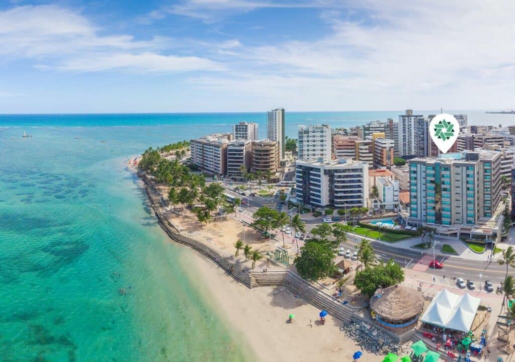 Vista aéra de uma parte costeira da orla em Maceió, onde tem um mar esverdeado transparente e, à direita, a orla com prédios e casas. Entre os prédios está o hotel Ponta Verde