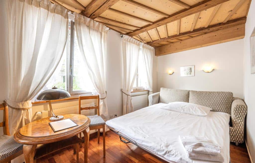 Foto do Hotel Rebstock, para representar o post de hotéis em Lucerna. O quarto possui um sofá-cama na direita, e uma mesa de madeira com abajur em cima e duas cadeiras está na sua frente, no canto esquerdo da foto. Há duas janelas no lado da cama que iluminam bastante a suíte.