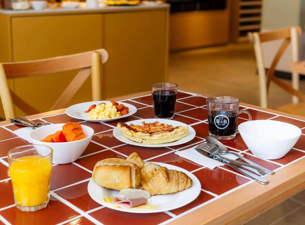 Mesa do café da manhã do B&B HOTEL Santos Dumont. Xícaras com café, um copo de suco, e pratos com pães, frios, talheres, frutas e salgados.