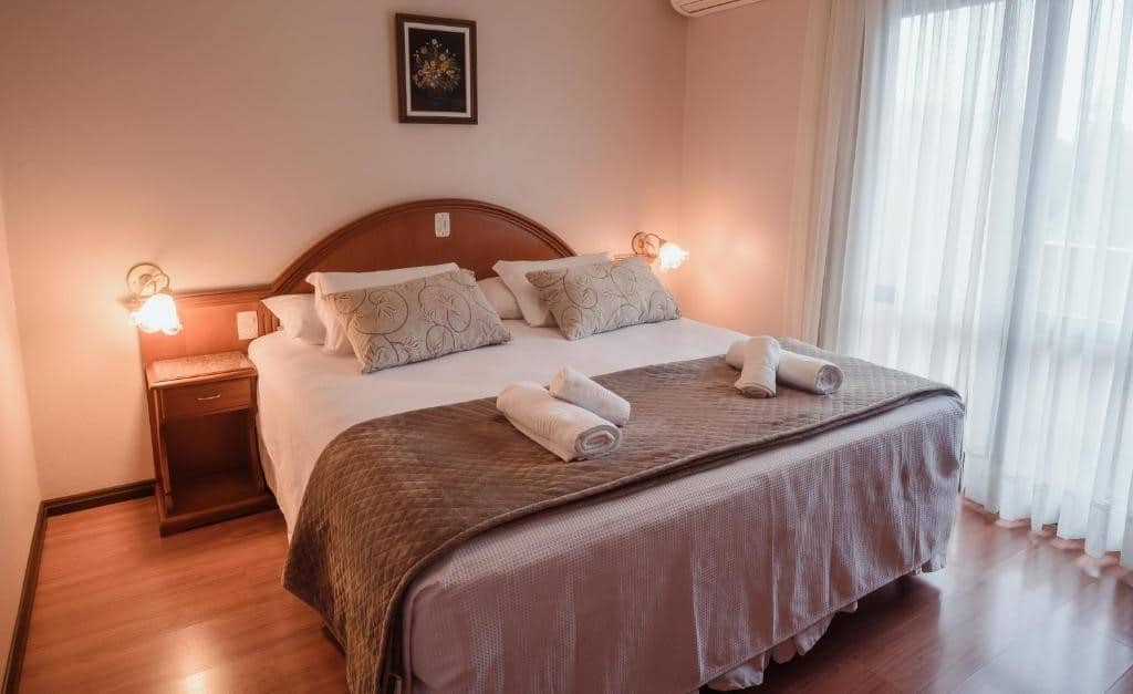 Foto do quarto do Hotel Vista do Vale para ilustrar post sobre hotéis baratos em Gramado. Uma cama de casal no meio com toalhas decorando, de cada lado uma cômoda com uma luminária. No lado direito a cortina fechando a janela do quarto.