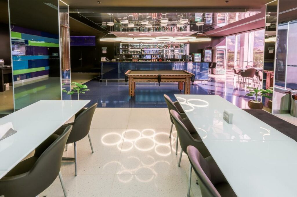 Área comum do hotel ibis Styles Confins Aeroporto. Duas mesas retangulares de vários lugares estão em primeiro plano enquanto ao fundo está o bar e uma mesa de bilhar.